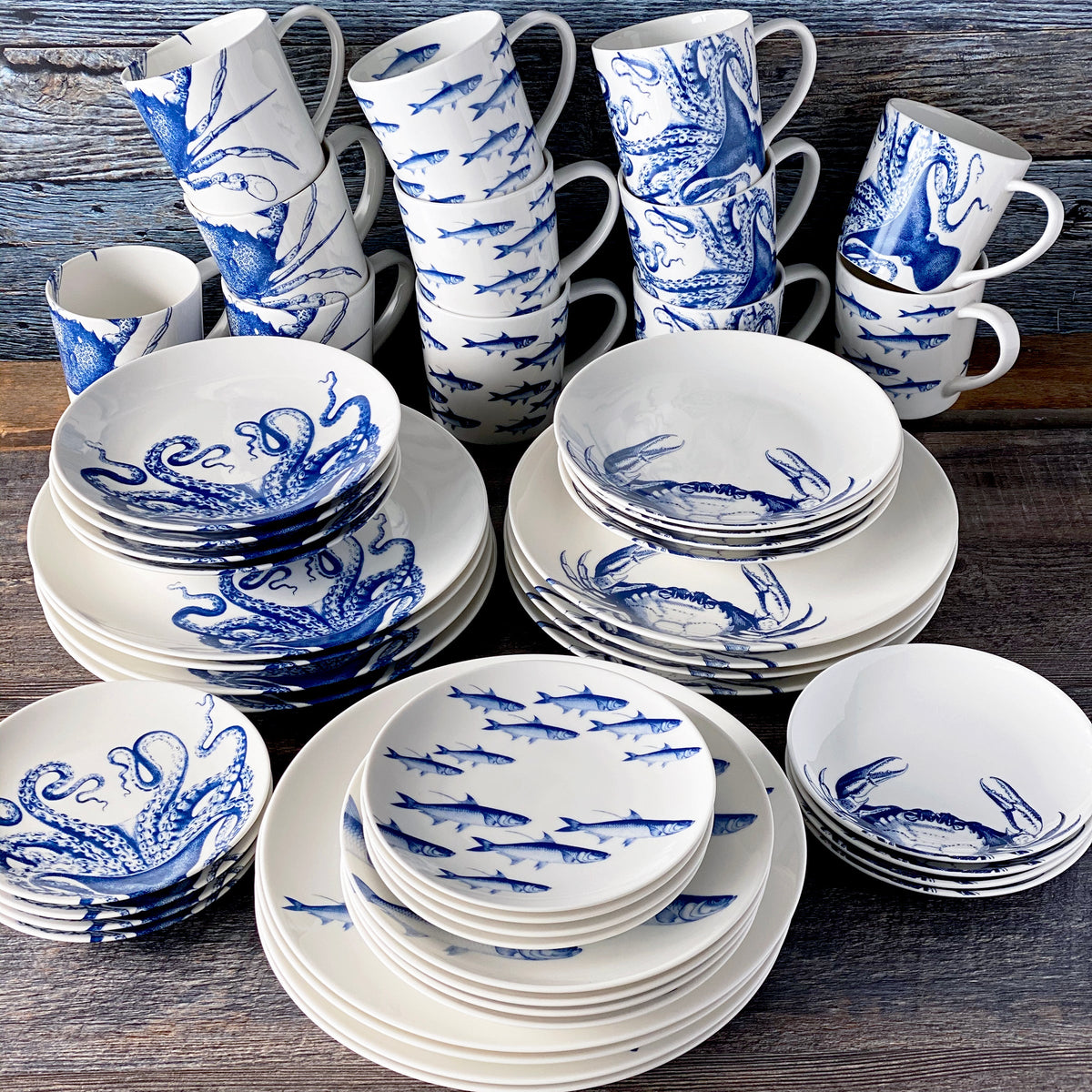 A Coastal Collection 48 Pc. Set by Caskata Artisanal Home, consisting of blue and white plates and cups, is placed on a wooden table, exuding a tranquil sea and ocean vibe with beautiful patterns.
