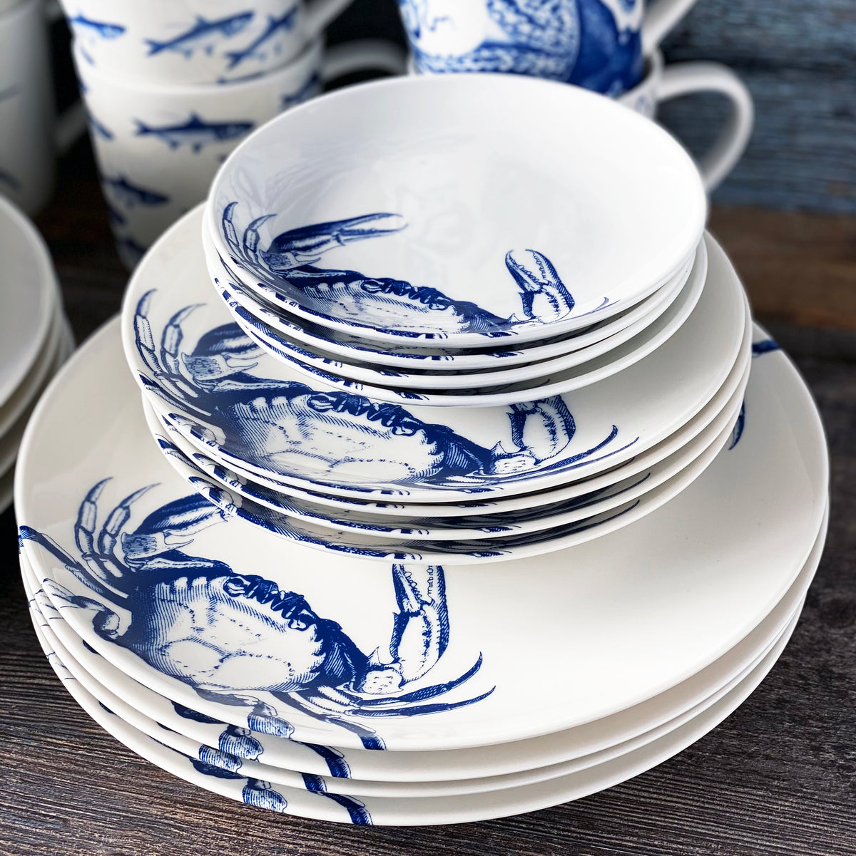 A stack of heirloom-quality dinnerware, including white ceramic bowls and Caskata Artisanal Home Crab Small Plates featuring a detailed crab pattern, is arranged on a wooden surface.