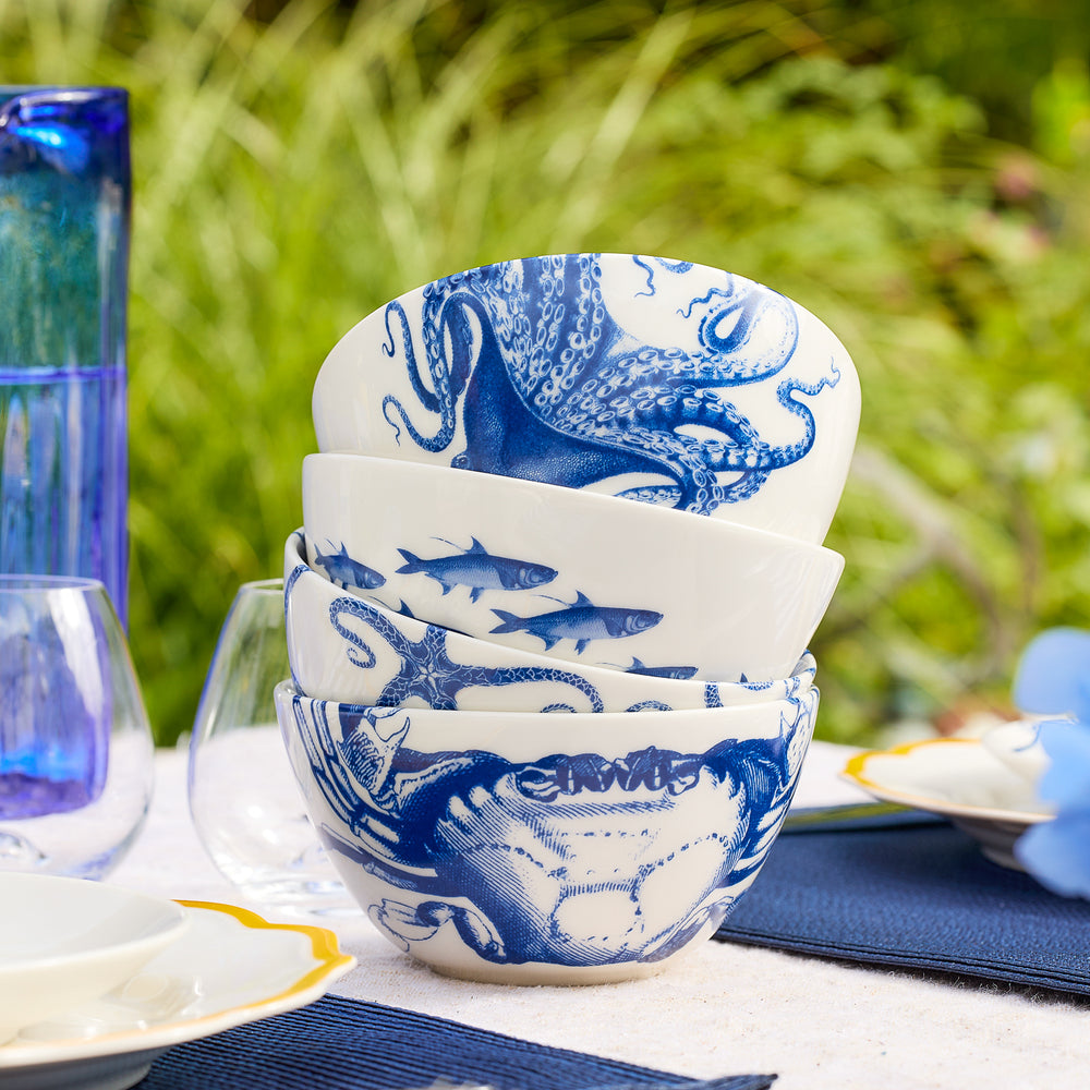 Four white porcelain Coastal Icons Cereal Bowls by Caskata, featuring blue marine life designs such as an octopus, fish, and crab, are stacked on an outdoor table set for a meal. This charming mixed set of four is perfect for seaside gatherings, and the dishwasher safe bowls make cleanup a breeze.
