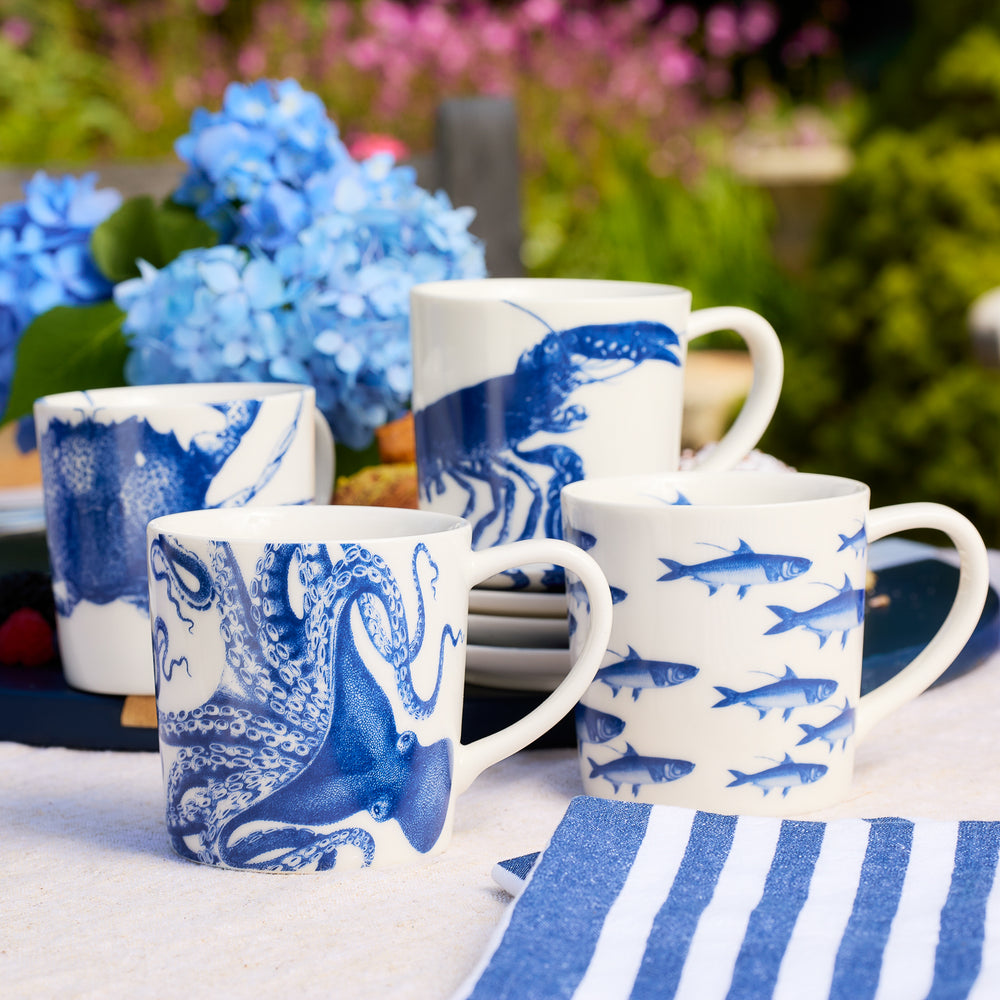 Four white porcelain mugs from Caskata’s Coastal Icons Mugs, Mixed Set of 4, featuring blue marine animal designs—octopus, lobster, and fish—are arranged on an outdoor table alongside a blue-striped napkin and blue hydrangeas. This charming set combines coastal style with practicality, as the dinnerware is dishwasher safe.