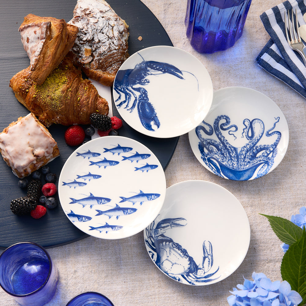 Caskata’s Coastal Icons Small Plates, Mixed Set of 4—featuring white porcelain with blue marine animal motifs—are beautifully arranged alongside pastries, berries, and blue glasses on a light tablecloth, creating a charming coastal-inspired breakfast or brunch setting.