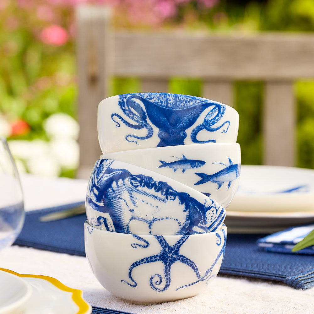 Four Caskata Coastal Icons Snack Bowls, a mixed set of four crafted from white porcelain and adorned with blue nautical designs—octopus, crab, and fish—are stacked on an outdoor dining table. This stylish dinnerware set is perfect for coastal-themed gatherings.