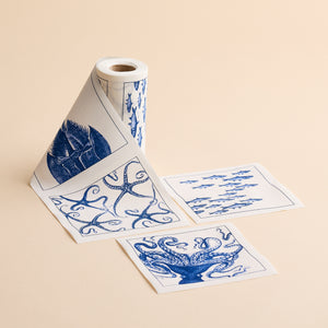 The MY DRAP Coastal Cocktail Napkin Roll features charming blue sea-themed designs, including fish, a starfish, and coral, unrolled on a beige surface—perfect for those seeking eco-friendly napkins with coastal patterns.