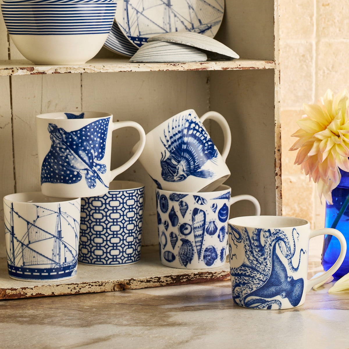 Six blue and white "Ray" the Stingray Mugs by Caskata, featuring nautical designs like fish, ships, seashells, and geometric patterns, are displayed on a shelf—perfect for any coastal mug collection.