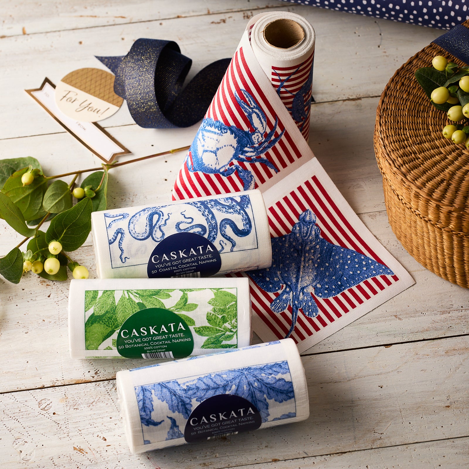Four MY DRAP Deluxe Entertainer Cocktail Napkin Rolls, featuring marine and plant patterns, are displayed on a wooden table near a woven basket and ribbon—ideal for stylish, reusable cocktail napkins.