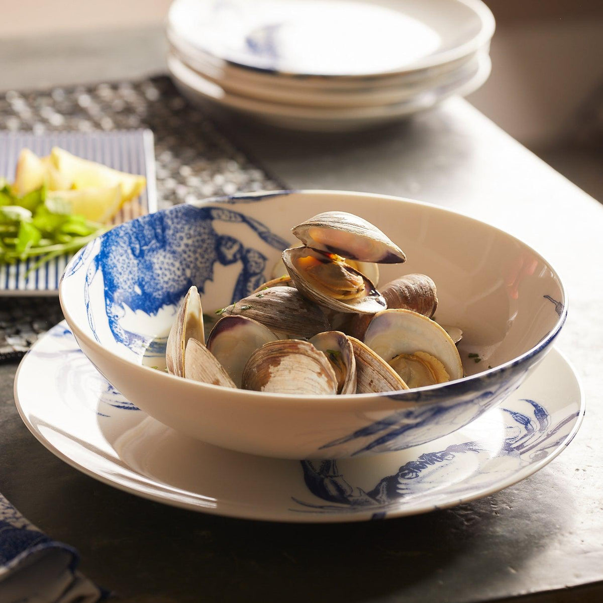 A **Crab Entrée Bowl** by **Caskata Artisanal Home** with blue designs and a crab pattern contains several clams. In the background, there is another plate of food and stacked white plates. Made of high-fired porcelain, it’s both dishwasher and microwave safe. The surface is dark-colored.