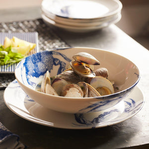 A **Crab Entrée Bowl** by **Caskata Artisanal Home** with blue designs and a crab pattern contains several clams. In the background, there is another plate of food and stacked white plates. Made of high-fired porcelain, it’s both dishwasher and microwave safe. The surface is dark-colored.