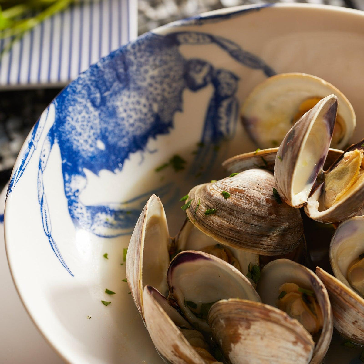 Bowl of cooked clams garnished with herbs, set in a dish adorned with a Crab pattern illustration on the inside. Made from high-fired porcelain, the Crab Entrée Bowl by Caskata Artisanal Home is both dishwasher and microwave safe. A striped linen cloth is partially visible in the background.