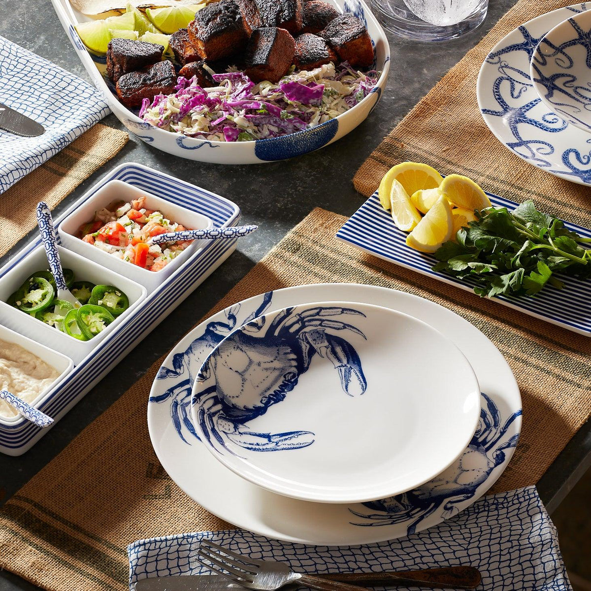 A dining table setting featuring premium Caskata Artisanal Home Crab Coupe Salad Plate with a blue crab pattern, a platter of food with charred items, side dishes including sliced peppers and a green herb, and a bowl with lime and lemon wedges.