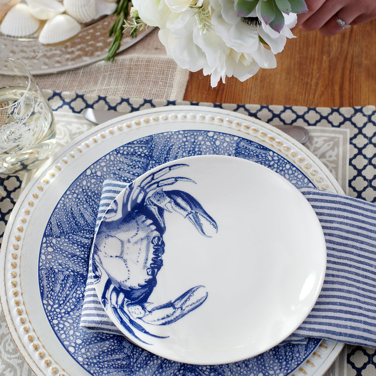 A table setting with heirloom-quality dinnerware boasts a navy and white Caskata Artisanal Home Crab Small Plates, placed on a striped napkin and larger plate. A glass of water, a bouquet of white flowers, and shells in the background complete the charming scene.
