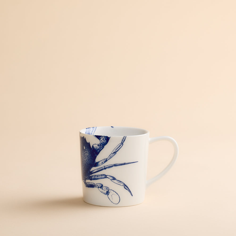 Caskata Artisanal Home’s Crab Mug—a white ceramic coastal mug adorned with a blue crab illustration—displayed on a simple beige background.