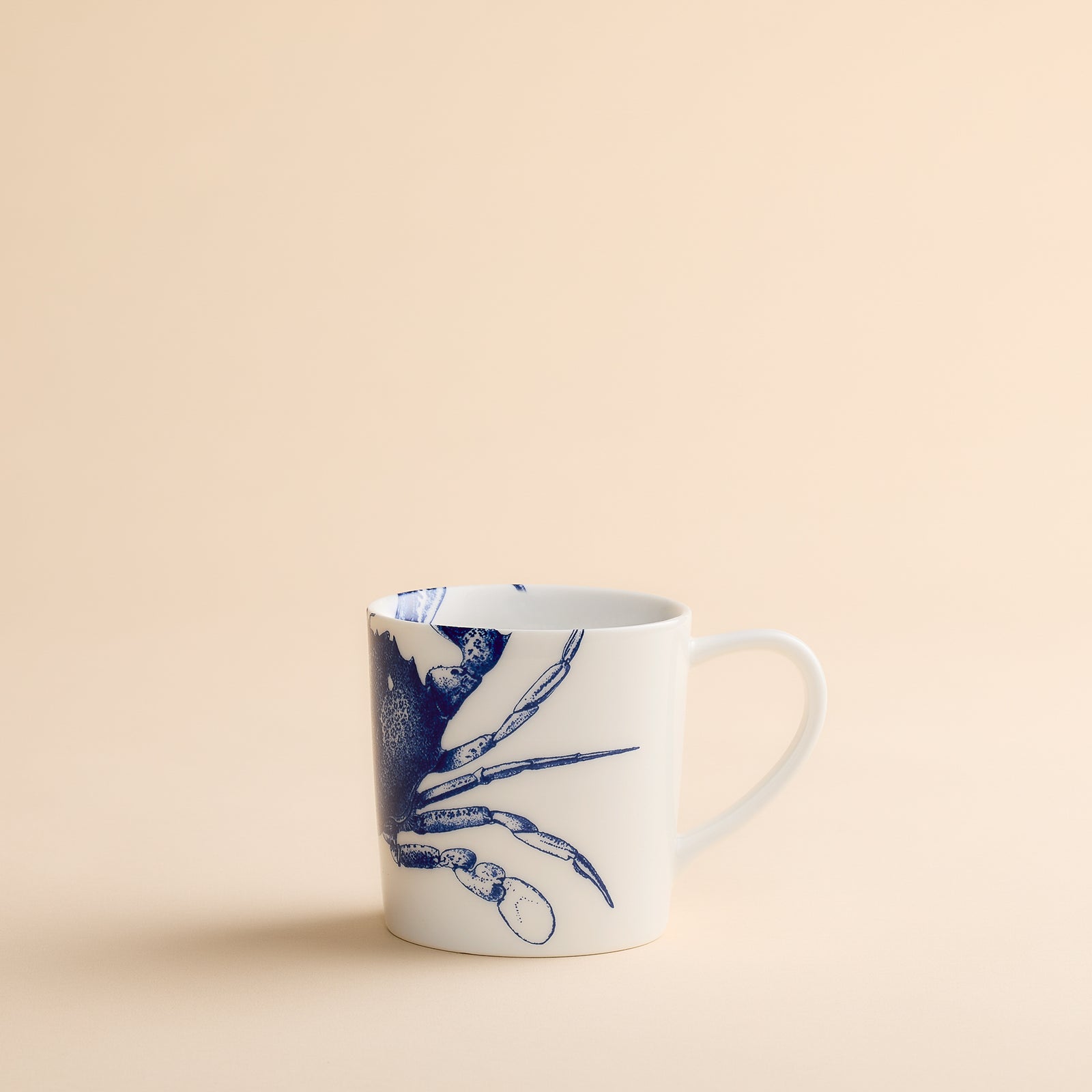 Caskata Artisanal Home’s Crab Mug—a white ceramic coastal mug adorned with a blue crab illustration—displayed on a simple beige background.