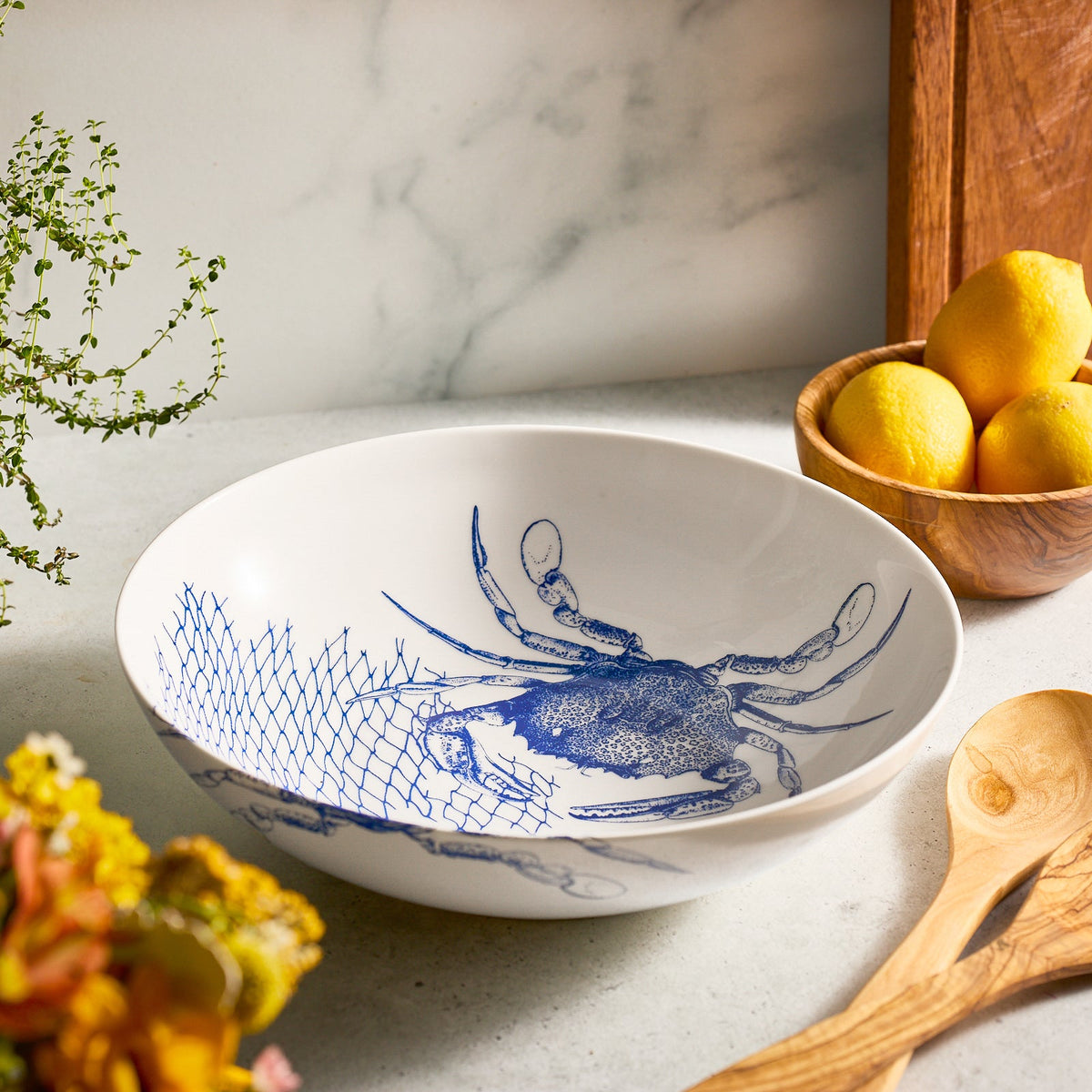 A Caskata Artisanal Home Crab Wide Serving Bowl, featuring a blue crab and net design, sits on the kitchen counter near a wooden bowl of lemons, a wooden spoon, and floral decor.
