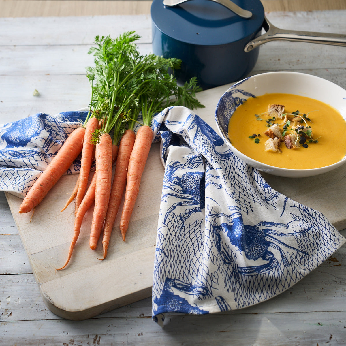 A bowl of soup and carrots on a cutting board with Caskata&#39;s Crabs &amp; Nets Kitchen Towels Set/2.