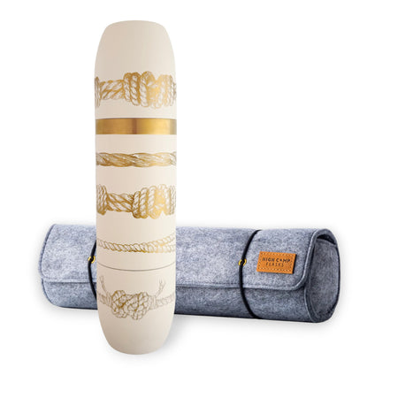 A cylindrical white vessel adorned with gold sailor's knot designs stands before a gray felt roll-up case labeled "High Camp Flasks." The Caskata x High Camp Crema Sailor's Knots Wine Flask and Case Bundle by Caskata features an insulated wine flask with magnetized cups, blending elegance and practicality for your next adventure.
