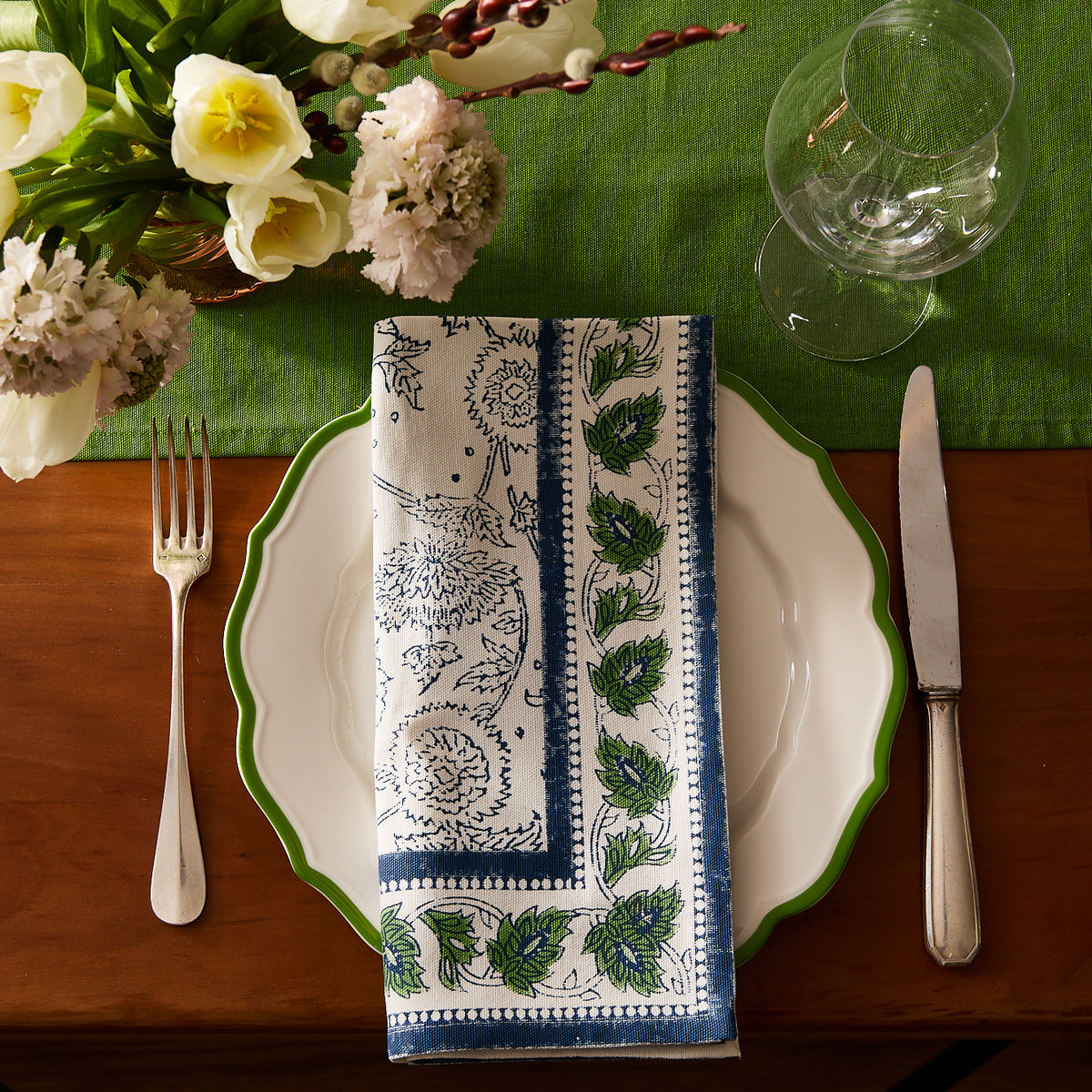 A table setting with a white plate, Caskata Cuttyhunk Block Print Dinner Napkin, fork, knife, wine glasses, and a green tablecloth, accented by a flower arrangement in the corner.