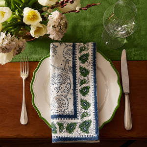 A table setting with a white plate, Caskata Cuttyhunk Block Print Dinner Napkin, fork, knife, wine glasses, and a green tablecloth, accented by a flower arrangement in the corner.
