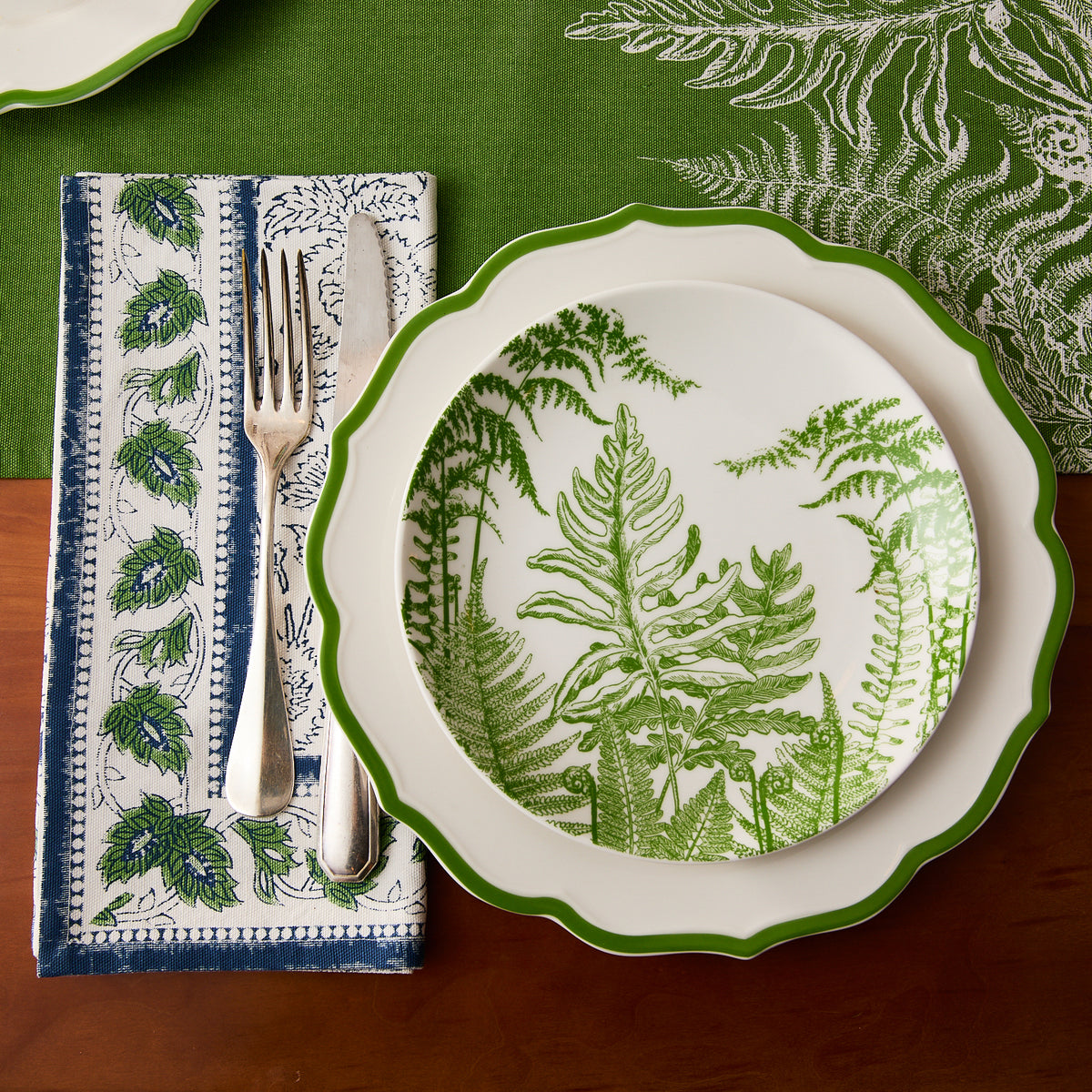 A dinner plate with a green fern pattern sits on a green tablecloth, with a fork and knife placed on Caskata's Cuttyhunk Block Print Dinner Napkins featuring leafy designs.