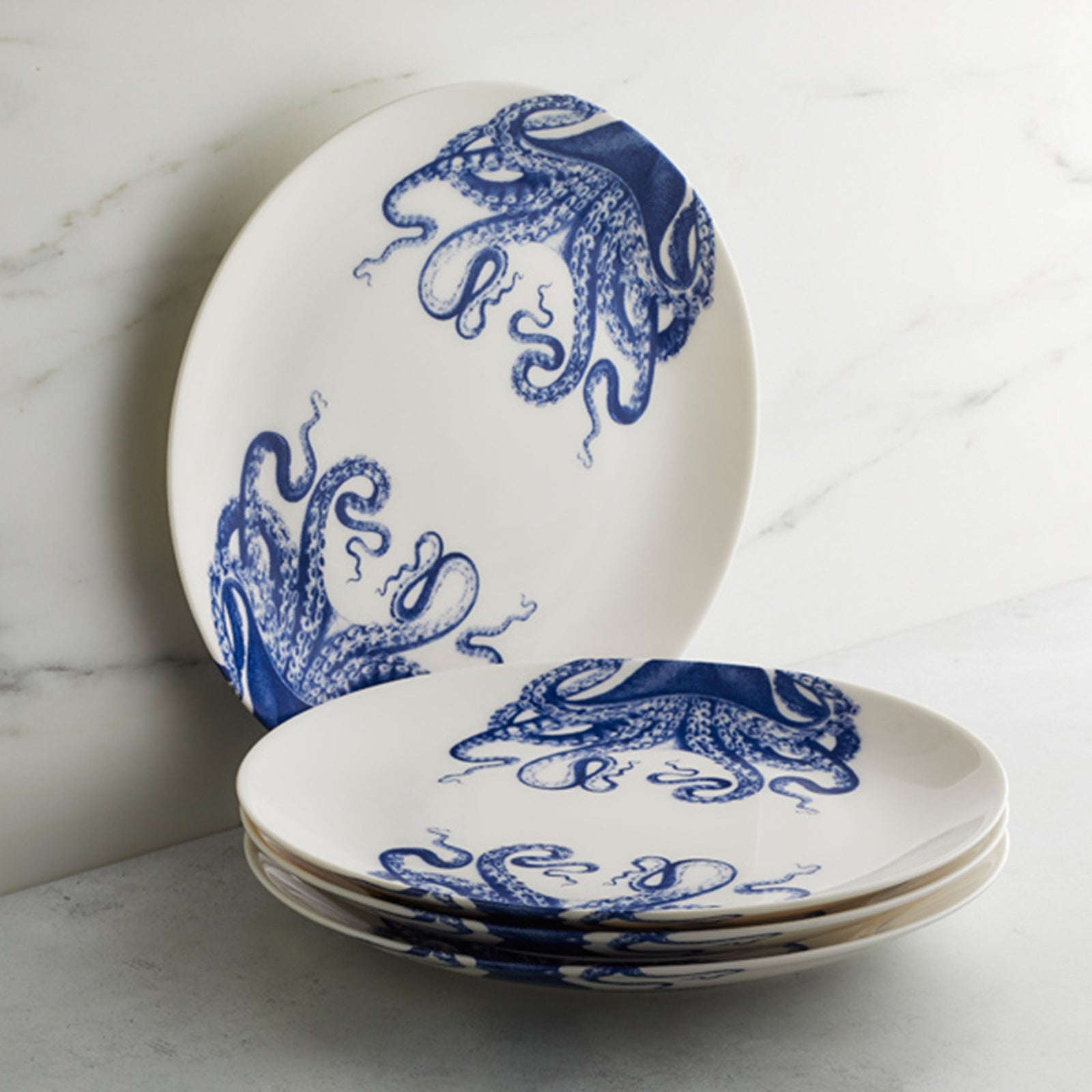 Four Caskata "Lucy Table for 4" plates feature elegant blue octopus designs. Three are stacked, with one upright behind them, beautifully displayed against a marble background.