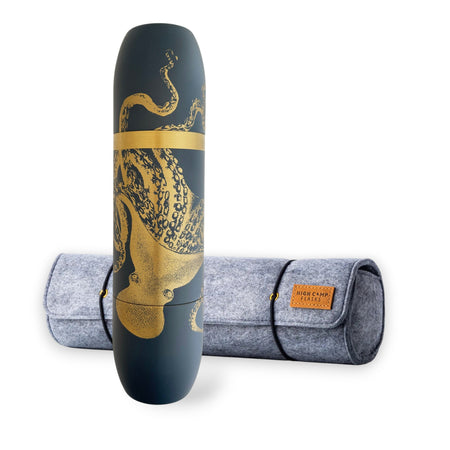 The Caskata X High Camp Flasks Lucy Dark Blue Flask stands upright, featuring a gold octopus design, in front of its gray felt case with a brown leather patch.