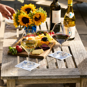 A wooden table set with Caskata Library White Wine Glasses (Set of 4), a cheese and fruit platter, two wine bottles, and a vase of sunflowers.