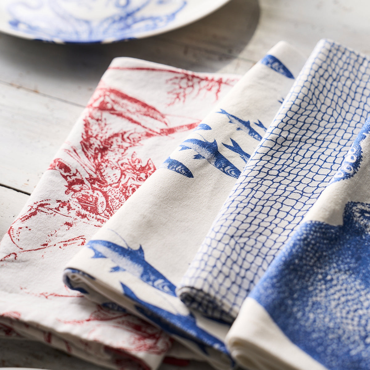 Four 100% cotton Catch Dinner Napkins, Set of 4 by Caskata with red and blue fish and sea creature prints are neatly arranged on a white wooden surface, featuring a charming coastal pattern, with a plate partially visible in the background.