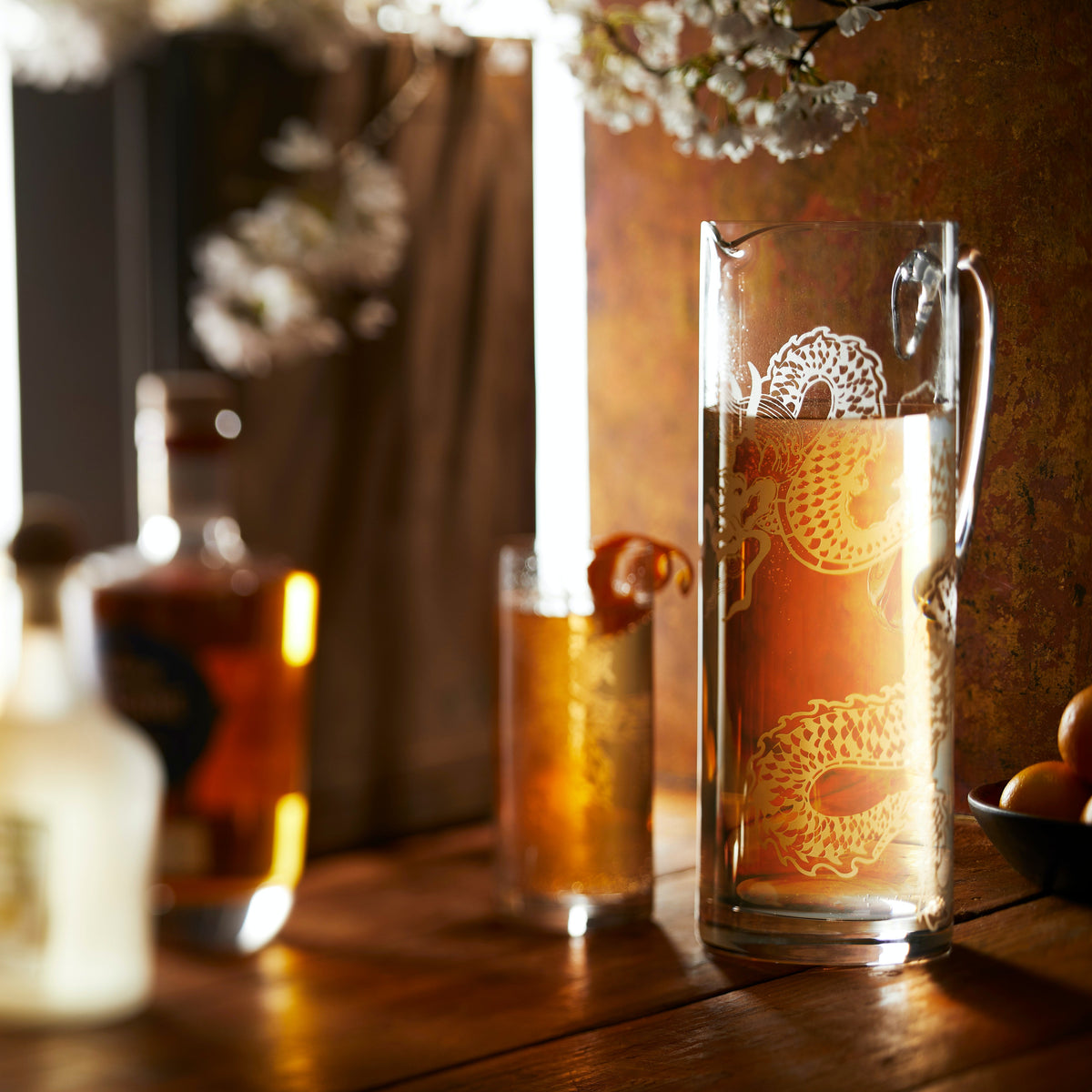 A sand etched dragon pitcher by Caskata filled with a cocktail on a rustic bar showcases the intricate design of the pattern.