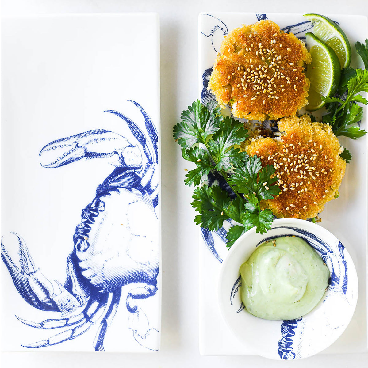 Two golden-brown crab cakes garnished with sesame seeds and cilantro, served with lime wedges and a small bowl of green dipping sauce on Caskata&#39;s Crab Medium Sushi Trays, Set of 2 featuring a chic crab pattern.