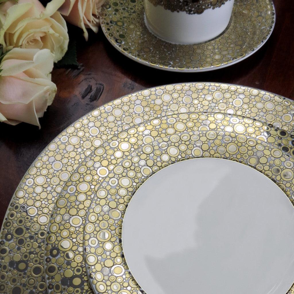 The Ellington Shimmer Gold & Platinum Bread & Butter Plate by Caskata Artisanal Home is a white ceramic plate adorned with a gold and beige bubble pattern on the rim. This hand-decorated bread plate brings an elegant harmony to any table setting and is dishwasher safe for easy cleaning.