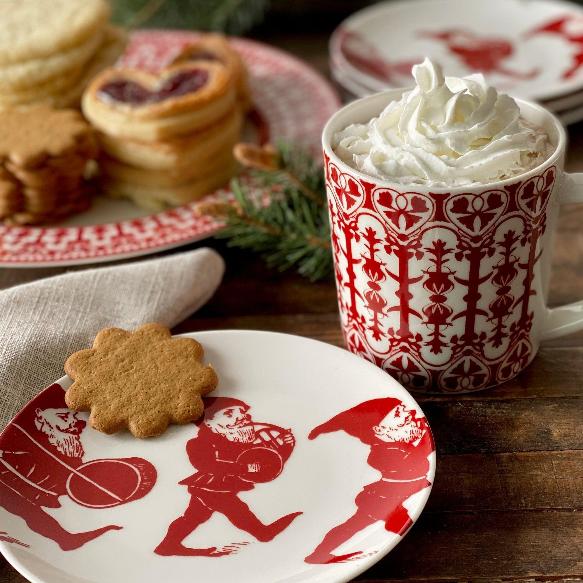 Four white plates, part of an heirloom-quality dinnerware holiday collection, each feature an illustration of a red gnome holding a drum. They are called Elves Small Plates by Caskata Artisanal Home.