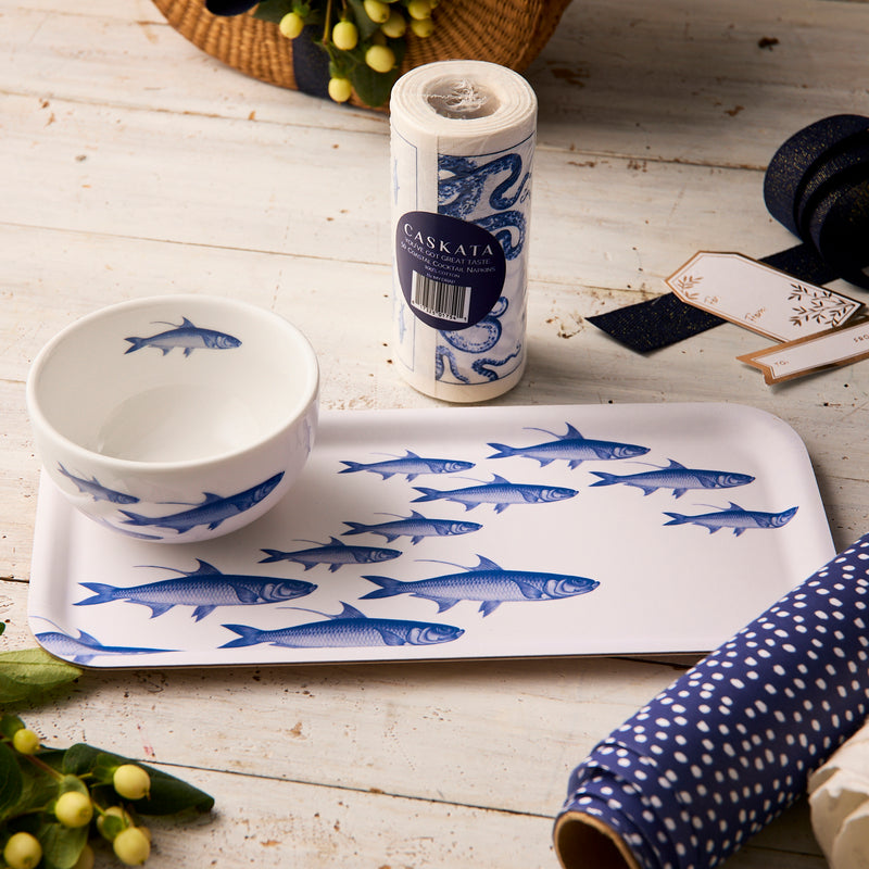 The Caskata "Everything's Going Swimmingly" Bundle, featuring a blue School of Fish appetizer tray and bowl set, is displayed on a wooden table with wrapping paper, ribbon, cocktail napkins, gift tags, flowers, and a wicker basket nearby.