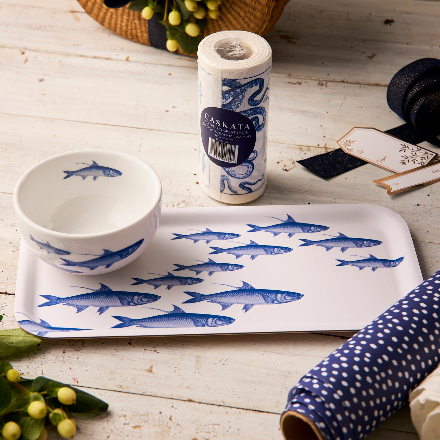 The Caskata "Everything's Going Swimmingly" Bundle, featuring a blue School of Fish appetizer tray and bowl set, is displayed on a wooden table with wrapping paper, ribbon, cocktail napkins, gift tags, flowers, and a wicker basket nearby.
