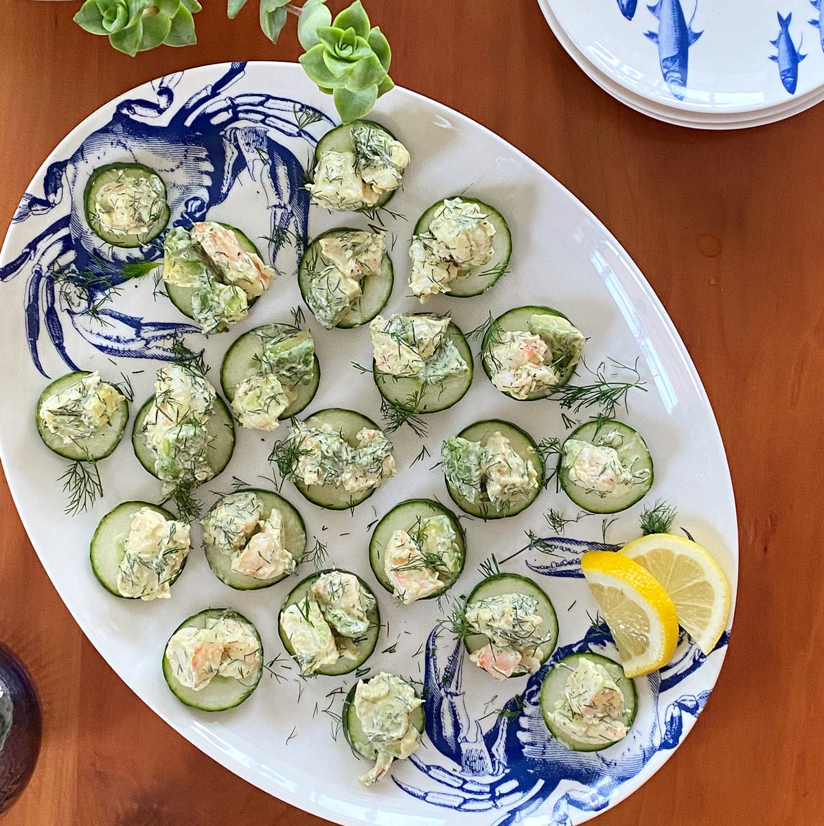 A Crab Coupe Oval Platter with cucumbers and lemon wedges on it, showcasing a coastal style. (Brand: Caskata Artisanal Home)