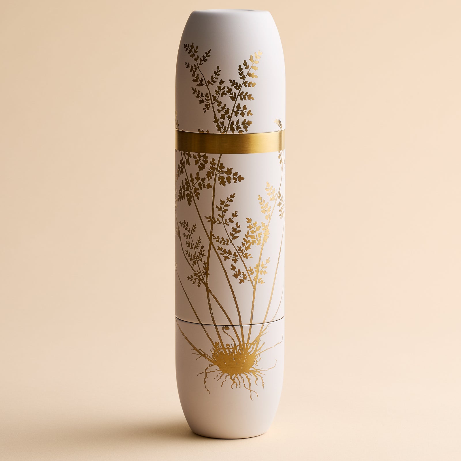 The Caskata x High Camp Crema Fairy Fern Wine Flask by Caskata, a tall white insulated bottle featuring gold botanical illustrations and a horizontal gold band around the middle, is showcased against a beige background.