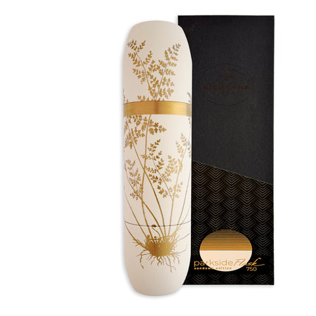 A white cylindrical Caskata x High Camp Crema Fairy Fern Wine Flask with a gold botanical design and band sits beside a black and gold box.