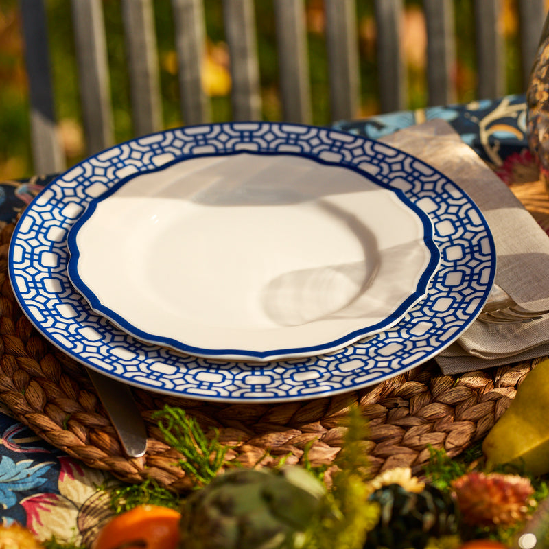 The Caskata Artisanal Home Newport Garden Gate Rimmed Dinner Plate, with its blue geometric border on white porcelain, is displayed on a woven placemat next to a napkin, knife, and autumnal table decor.