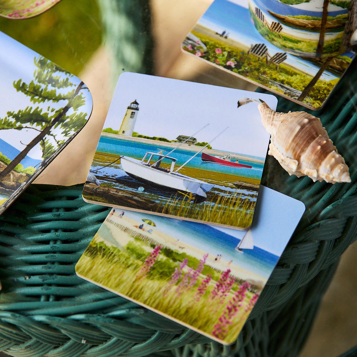 Caskata x Felix Doolittle Summer Birchwood Coasters, Set of 4, featuring beach and boat scenes, rest on a green wicker table beside a seashell, reflecting the coastal charm of Caskata’s birchwood designs.