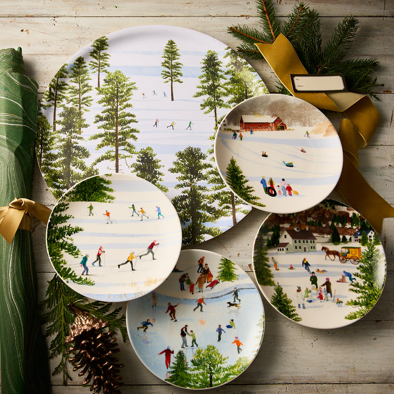 Caskata’s "Felix's Frolic" Bundle includes five decorative plates with winter sports scenes, artfully displayed on wood with pine, ribbon, and a green napkin—ideal for festive holiday entertaining and set displays.