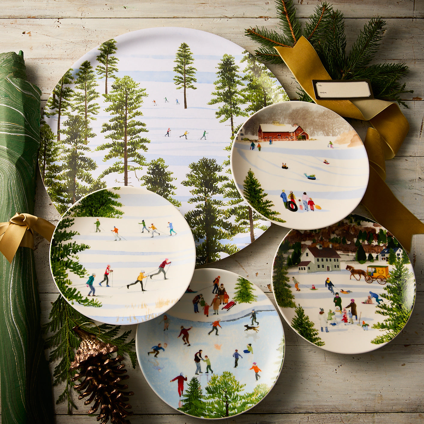 Caskata’s "Felix's Frolic" Bundle includes five decorative plates with winter sports scenes, artfully displayed on wood with pine, ribbon, and a green napkin—ideal for festive holiday entertaining and set displays.