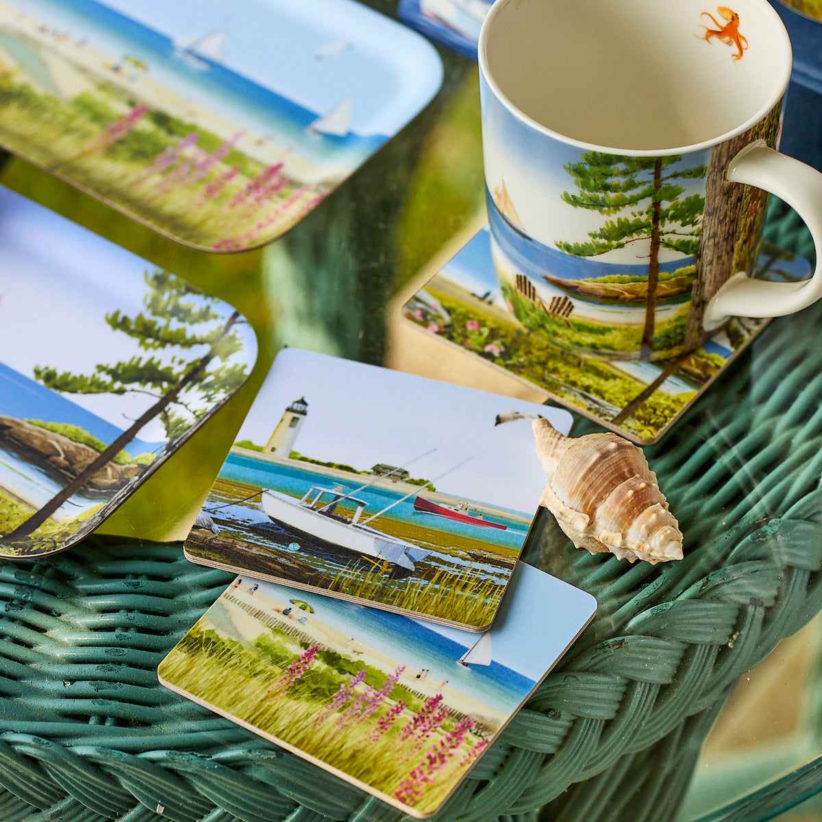 Caskata X Felix Doolittle Summer Birchwood Coasters, Set of 4, are arranged with mugs and plates on a green wicker table beside a seashell, adding eco-friendly elegance and coastal charm to your décor.