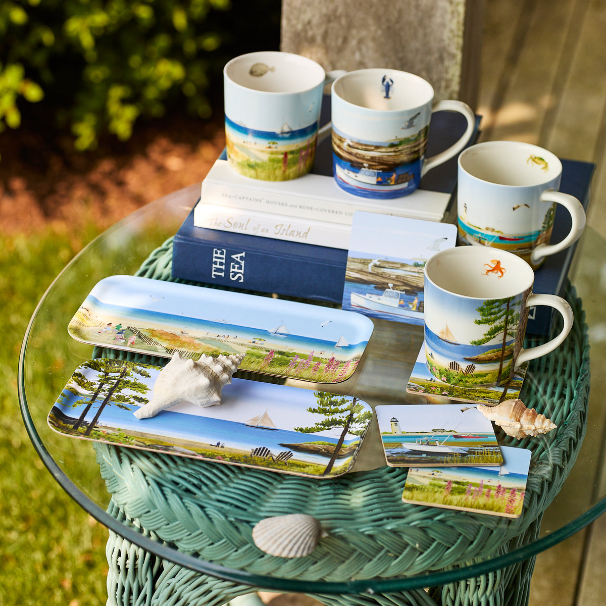 A glass table displays Caskata X Felix Doolittle Beach Dunes Small Rectangular Tray, mugs, and plates decorated with coastal scenes and seashells—the perfect summer home decor set outdoors on a wicker base.