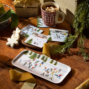 A festive mug of hot chocolate with marshmallows sits on Caskata's Snowball Fun Small Rectangular Tray, atop a wooden table surrounded by a gift box, ribbon, and pine branches—a cozy, sustainable gift idea.
