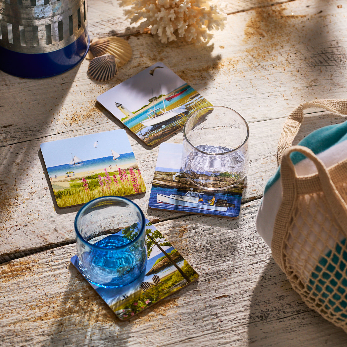Two glasses of water rest on Caskata X Felix Doolittle Summer Birchwood Coasters, set of 4, their beach-inspired designs adding relaxed New England summer charm to a rustic table decorated with seashells and a striped bag.
