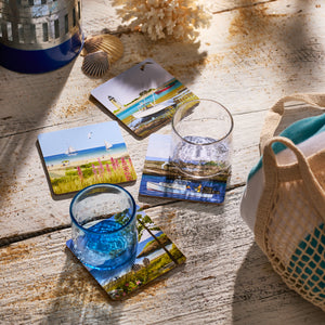 Two glasses of water rest on Caskata X Felix Doolittle Summer Birchwood Coasters, set of 4, their beach-inspired designs adding relaxed New England summer charm to a rustic table decorated with seashells and a striped bag.