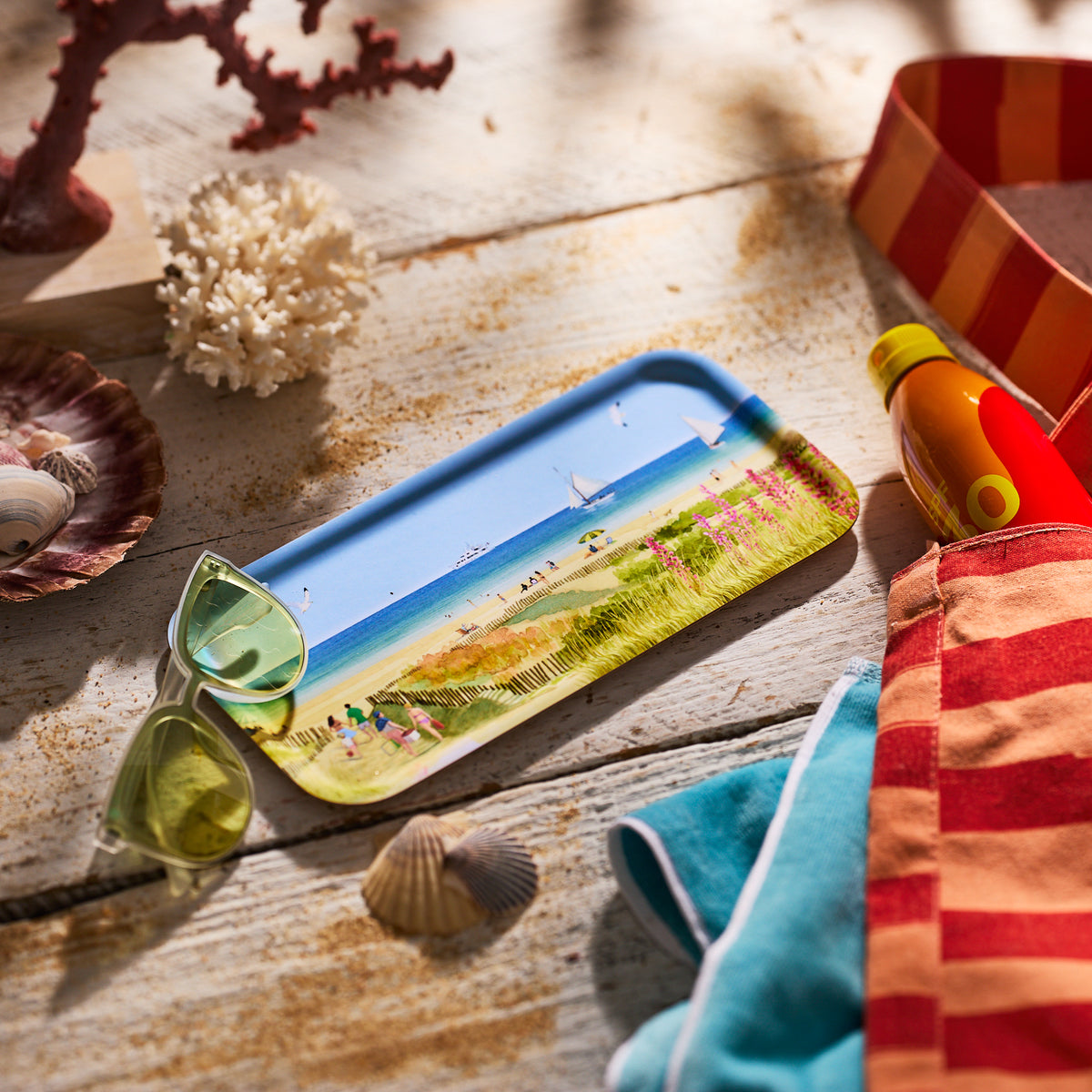 The Caskata X Felix Doolittle Beach Dunes Small Rectangular Tray by Caskata is styled with green sunglasses, a striped bag, towel, sunscreen, shells, and coral on sand-hued wood—ideal for summer home decor.