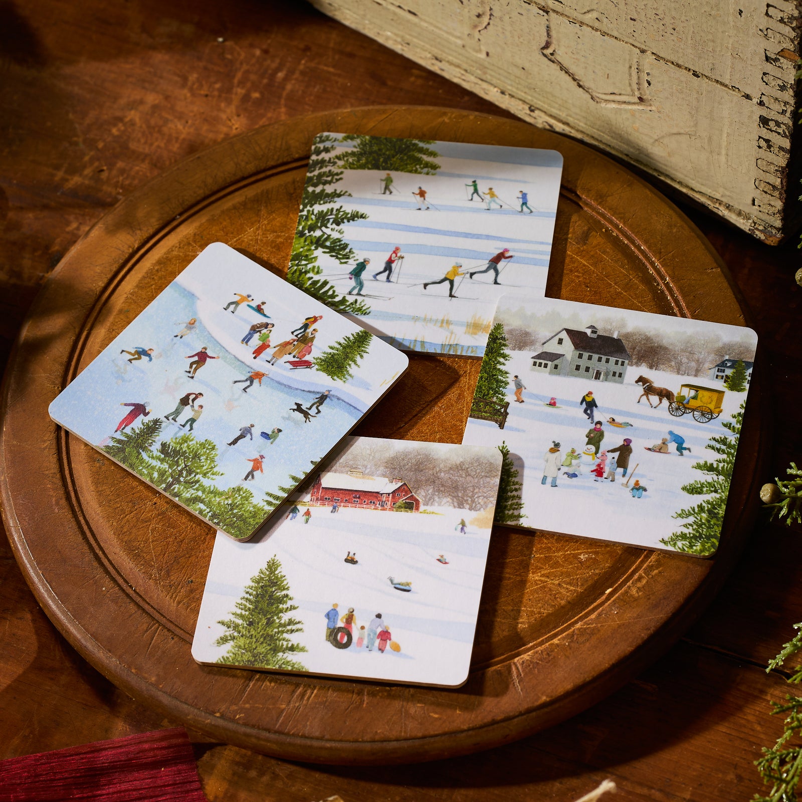 Four winter scenes—skiing, skating, sledding by a red barn, and a horse-drawn sleigh—decorate Caskata X Felix Doolittle Winter Birchwood Coasters. Set of 4 by Caskata; made in Finland from FSC-certified birchwood.