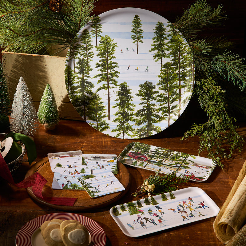 The Caskata X Felix Doolittle Winter Skiers Large Round Tray by Caskata is arranged on a wooden table with cookies, napkins, and festive decor. The tray features winter scenes and many pieces in the collection are made in Finland.