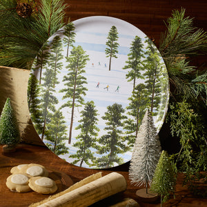 The Caskata X Felix Doolittle Winter Skiers Large Round Tray by Caskata showcases a winter forest with skiers, pine branches, cookies, and wooden props on a wooden surface—perfect as a decorative melamine tray.