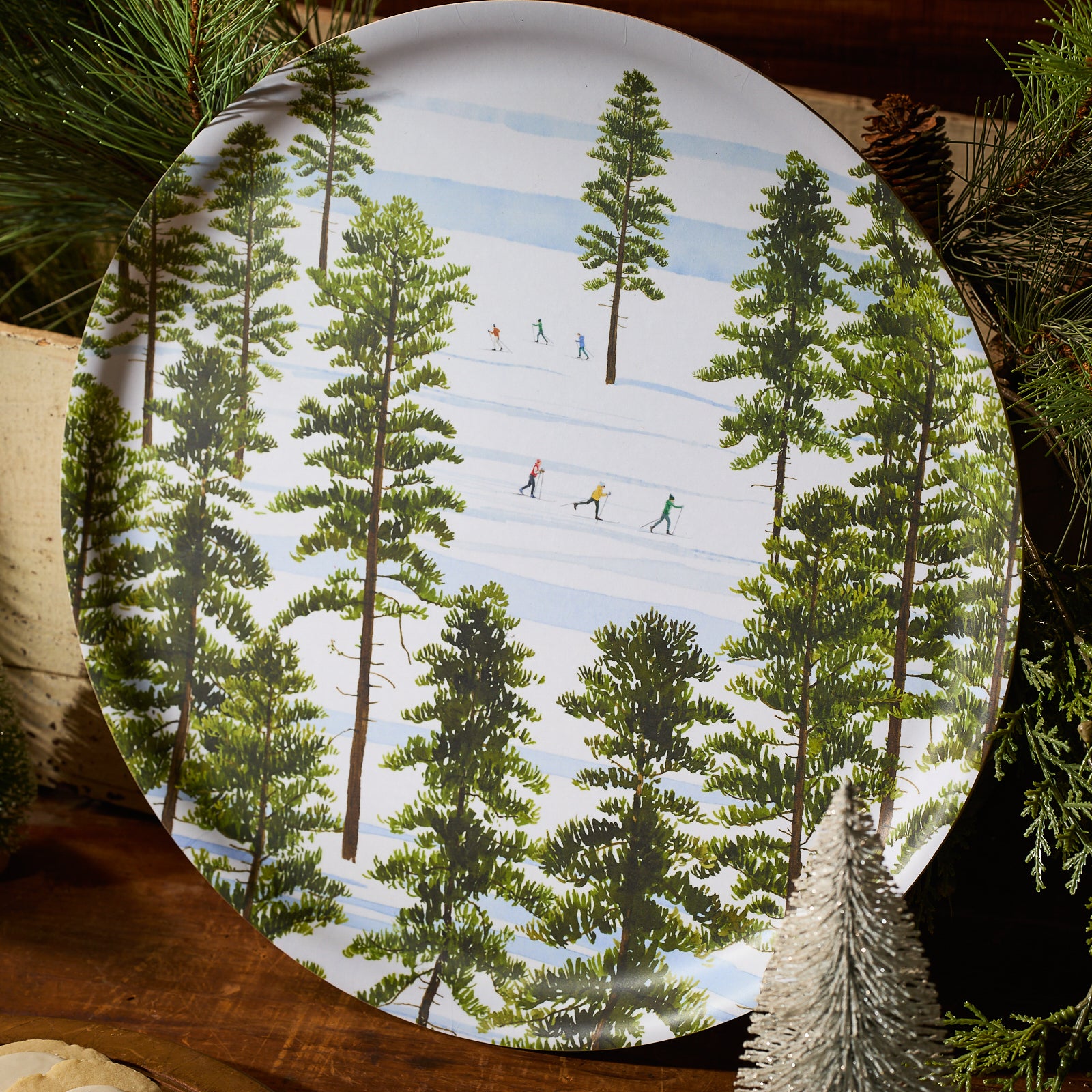 An overhead view of snowy evergreens and cross-country skiers inspires the Caskata X Felix Doolittle Winter Skiers Large Round Tray by Caskata, crafted from FSC-certified birchwood in Finland.