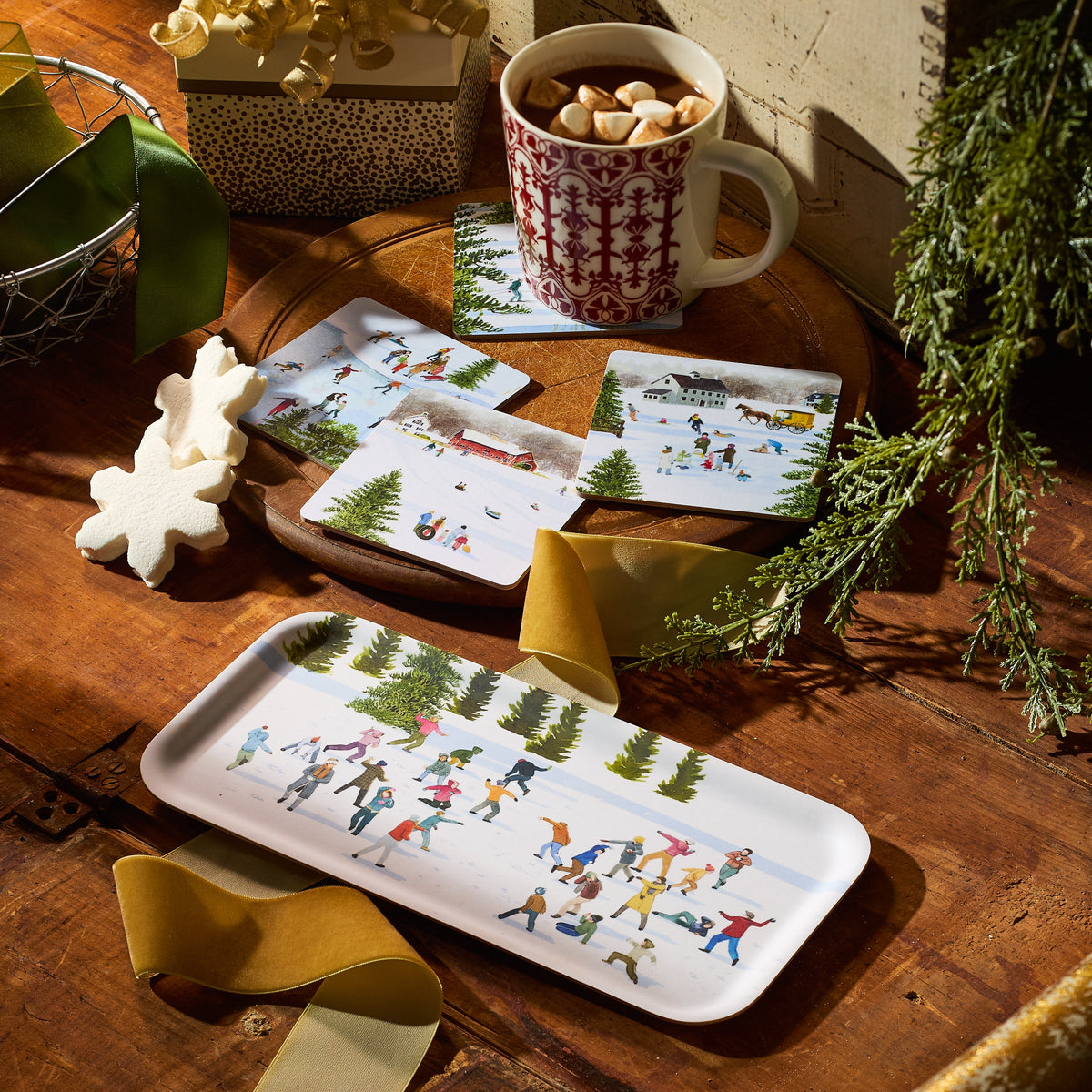 A set of four Caskata X Felix Doolittle Winter Birchwood Coasters, a mug of hot chocolate with marshmallows, ribbon, and snowflake cookies rest on a wooden table with evergreen branches. Made in Finland.